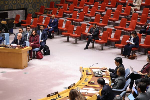 Diplomats sit at a table during a U.N. Security Council meeting. Mostly empty red chairs for audience members are nearby.