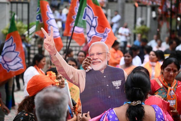 A crowd of people; a woman with her back to the camera is holding a life-size cutout of Prime Minister Modi making the “V” sign.