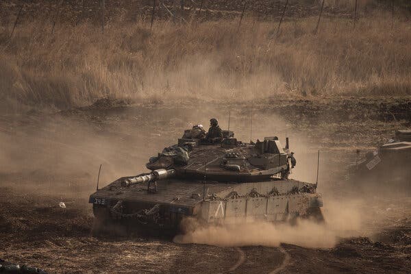 A figure in military gear rides atop a tank on dusty terrain.