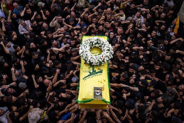 A coffin draped in a yellow and green cloth with a wreath atop it, being lifted over a crwod of men raising their hands.