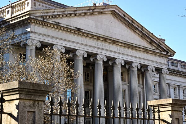 A large building with columns and the words "The Treasury Department" carved into the top, behind an iron fence with a blue sky in the background.