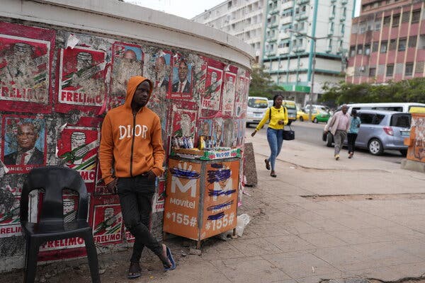 A man in an orange hoodie leans against a curved city wall checkered with peeling campaign posters. 