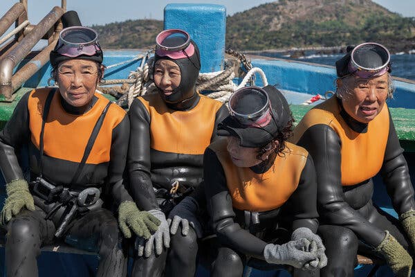 In a scene from the movie, four older woman in full orange-and-black wet suits, face masks raised, sit next to one another outdoors. 