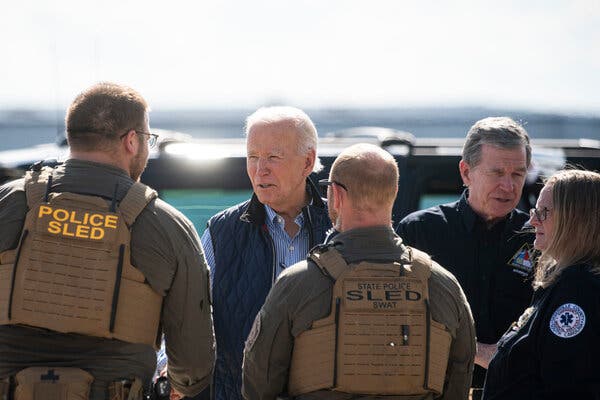 A person in a blue vest and a person in a black jacket talk to people in law enforcement wearing khaki green shirts and tan vests.