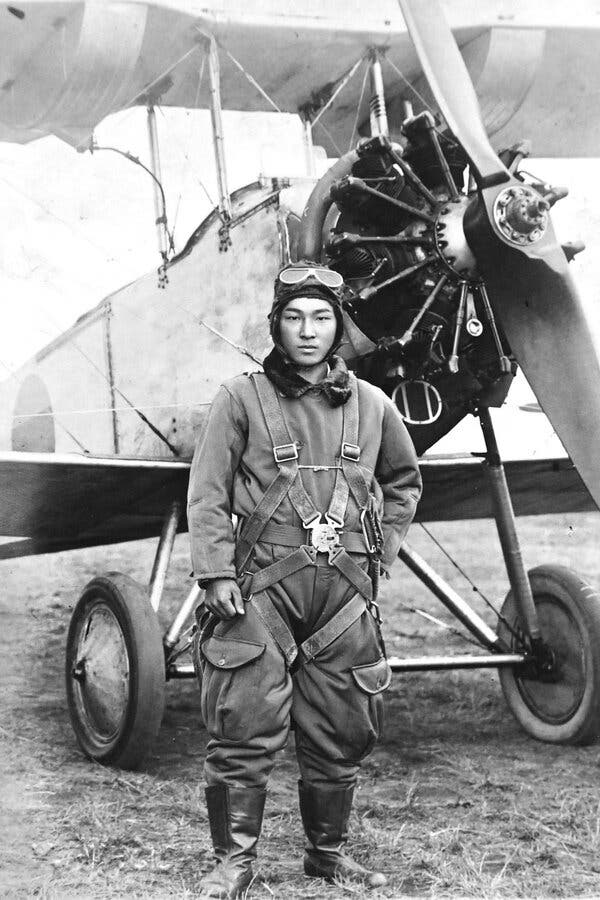 A black and white photo of Masamitsu Yoshioka wearing his pilot's overalls, his goggles atop his head, while posing in front of a small airplane.