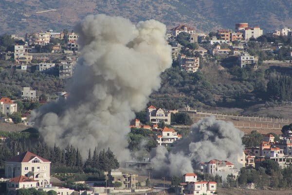 Two plumes of thick gray smoke billow above buildings and trees in the daytime.