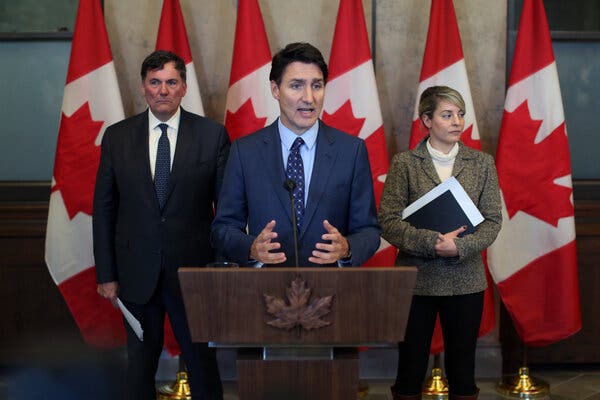 Prime Minister Justin Trudeau of Canada, in a blue suit and tie, stands at a podium with two officials behind him, as well as a row of Canadian flags.