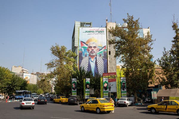 Cars drive on a street in front of a mural of Yahya Sinwar on the side of a building.