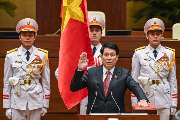 A Vietnamese politician, in a dark suit, raises his right hand while placing his left hand of a red book. Behind him are three people in military uniforms. One is holding a flag. 