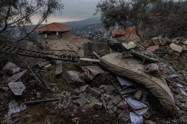 A large soiled mattress is piled atop other debris outside in the mud near spindly trees and a building.