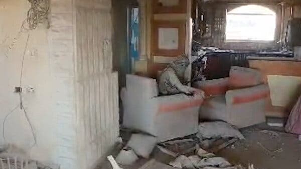 A video still showing a man, his head wrapped in a kaffiyeh and his clothes covered with dust, sitting in an armchair in a room visibly damaged by the war, with what look like sandbags on the floor. 