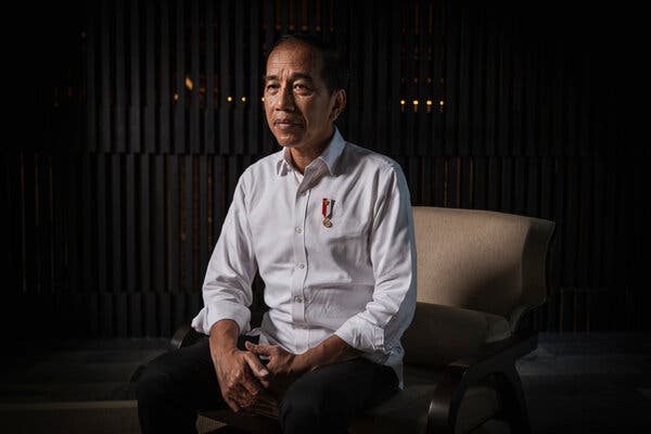 A man in white shirt and dark pants sitting on a chair. He has a medal of sorts pinned to his chest.