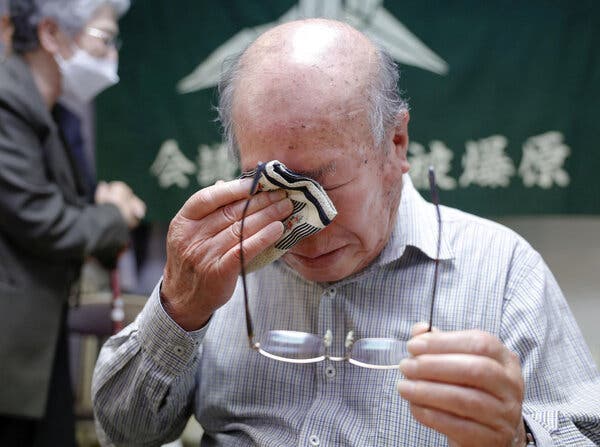 An older man holds spectacles in one hand and dabs at tears with a handkerchief with the other.