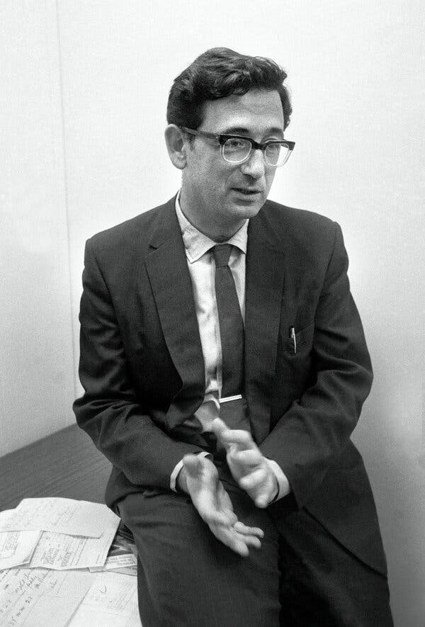 A young Yehuda Bauer sits on the edge of a desk, wearing glasses, a dark suit, a white shirt and a dark tie with a tie clip.