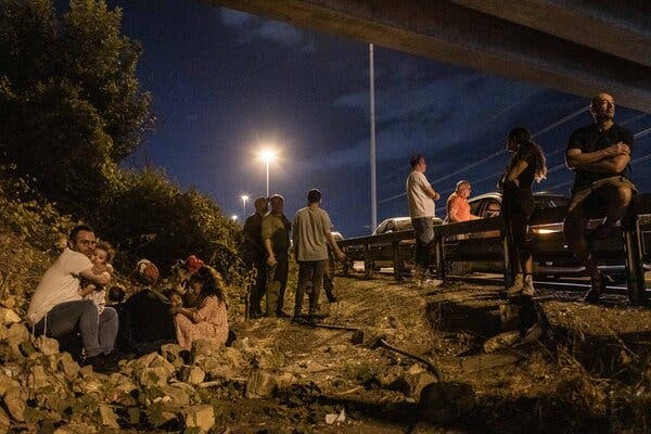 People standing and sitting by the side of a road at night; on the left it looks as if a family is sitting among rocks, with the man holding a baby tightly.  