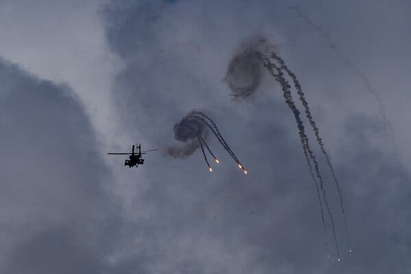 A helicopter is seen in shadow, with smoke and flares in the sky next to it.