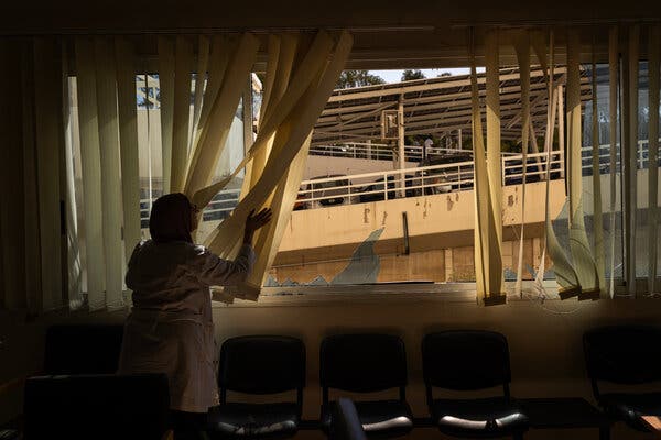 A nurse, in shadow, moves window covering from a damaged building,