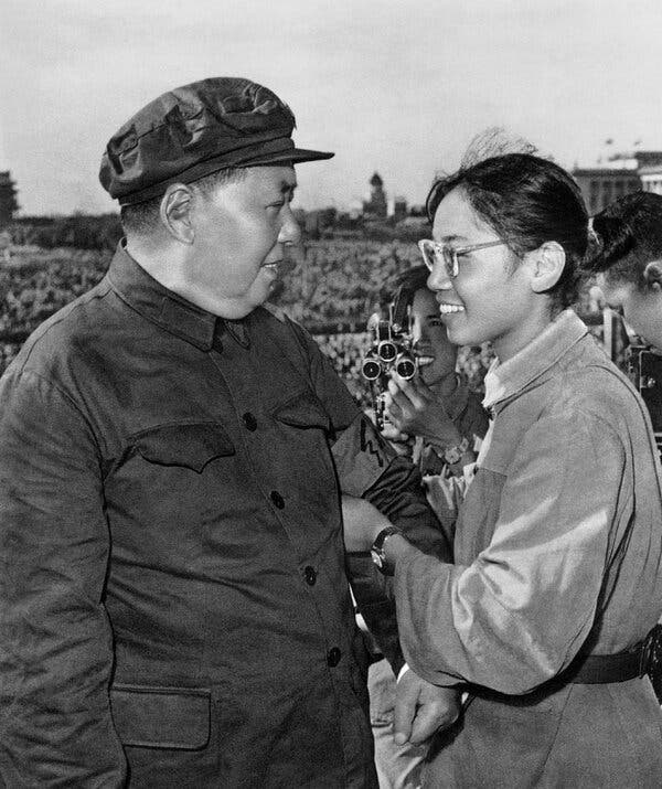 A black and white photo of Mao and a young woman smiling at each other as she places an armband around his sleeve. Behind them a young man films the event with a hand-held camera. Glimpses of a crowd can be seen below.