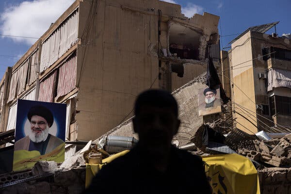 A man stands with his back to the camera, facing two posters with the image of the Hezbollah leader Hassan Nasrallah, amid buildings damaged by airstrikes.