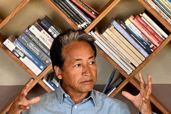 A man gestures with his hands in front of a slanted bookshelf.