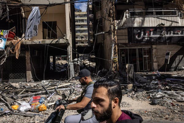 Two men on scooters ride down a street filled with rubble and demolished storefronts. 