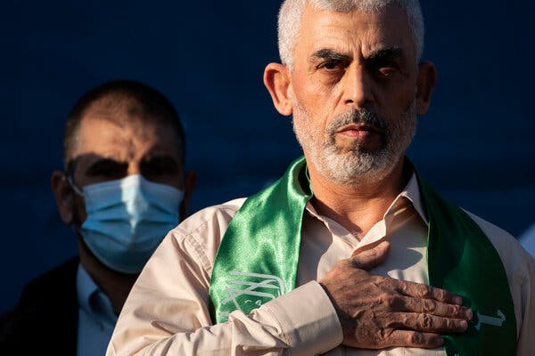 Yahya Sinwar, the leader of Hamas, is shown with his hand over his heart while standing on a stage in 2021 during a rally. A man wearing a medical mask stands behind him. 