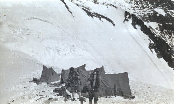 In a black-and-white image, two men stand by tents with a view of the snow-covered mountain.