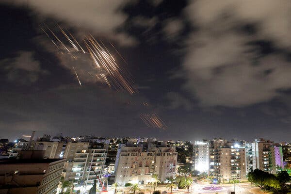 Streaks of light are crossing the night sky, over a city filled with tall buildings, many of which have their lights on.