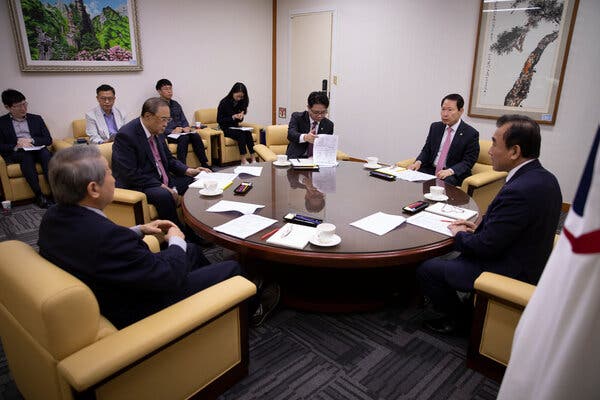 Five men in dark suits sit at a round table, each with a set of papers.  A few other people sit in chairs along a wall.