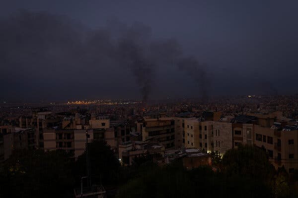 In the night sky, plumes of smoke rise from a settled area beyond some high-rise buildings. 