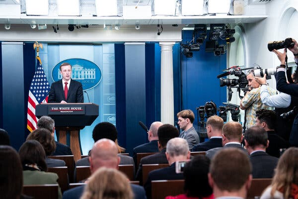 Jake Sullivan, in a blue suit and a red tie, stands behind a lectern facing a cluster of photographers and reporters