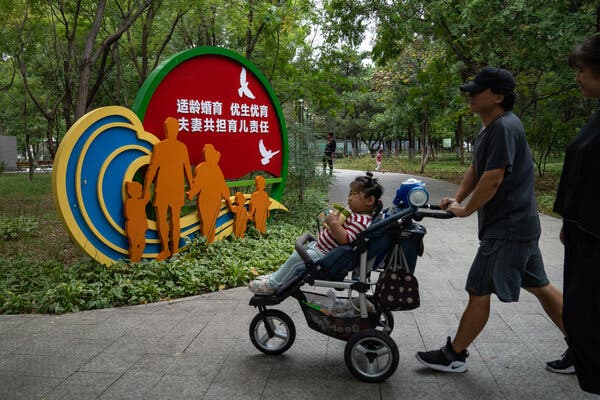 In a park, a family walks past artwork featuring life-size cutout of a man and woman walking with three children, under a slogan urging couples not to wait too long to have children.