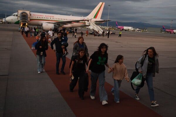 Families and others walking along a red path on an airport tarmac after leaving a small plane from the front.