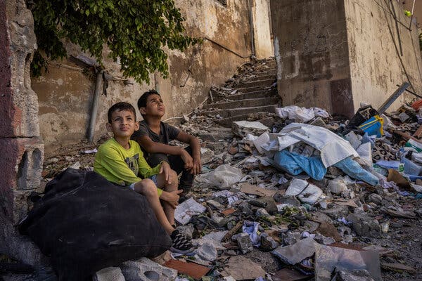 Two boys sit on the ground outside eyeing the sky while surrounded by debris beside a damaged building.