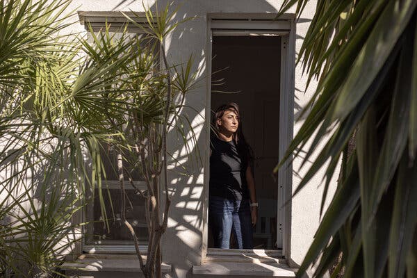 A woman in a black shirt and blue jeans looks out of a doorway that is bordered by a few trees.