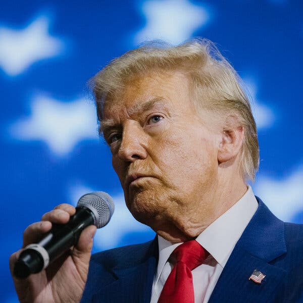 Donald Trump holds a microphone in his right hand, with white stars on a blue backdrop behind him.