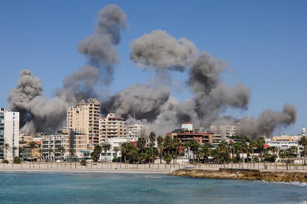 Plumes of thick smoke rise behind multistory buildings along a shoreline.