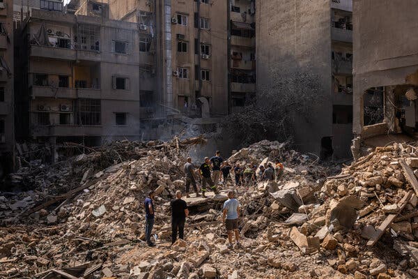 People stand on big piles of rubble near multilevel buildings.