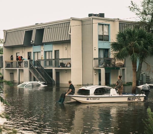 An apartment complex surrounded by water  with rescuers on a nearby boat. 