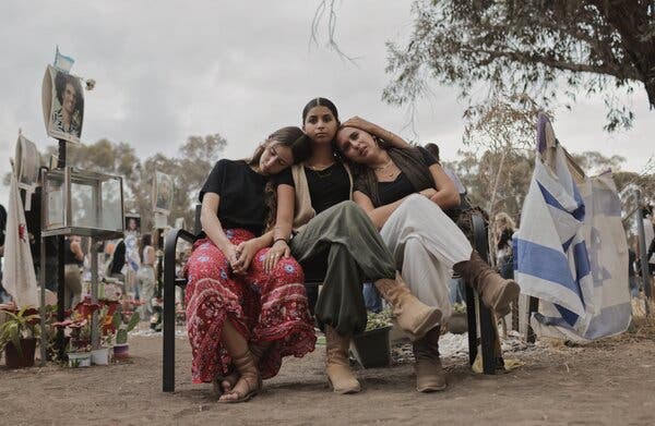 Three women leaning against one another on a bench, with flags and memorials around them and a crowd in the background.