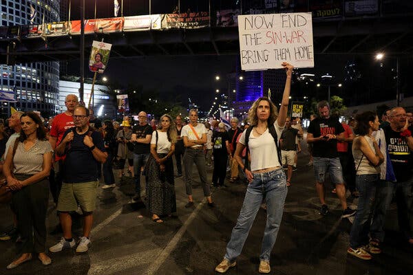 A group of people stands outdoors at night, some holding up signs, including a woman in the foreground whose sign says “Now end the sin-war bring them home.”