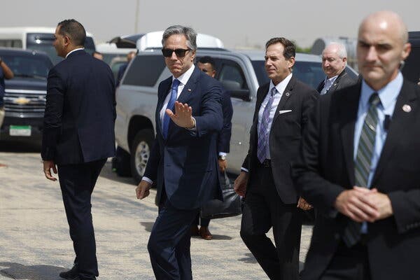 A man in a dark blue suit walks past parked cars with other men in suits. 