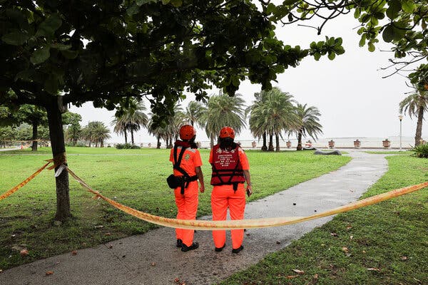 Two people wearing orange overalls and red helmets stand on a path that is blocked by warning tape. Trees dot the coastline in the background.