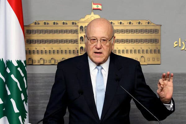 Najib Mikati speaks into a microphone, holding up one hand. A flag is off to the side. 