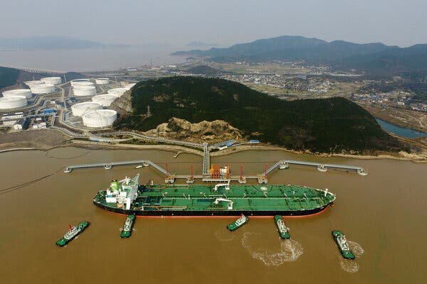 A long, wide oil tanker with a green deck and black sides is being pushed against an offshore network of pipes by three green tugboats as two more tugboats wait.