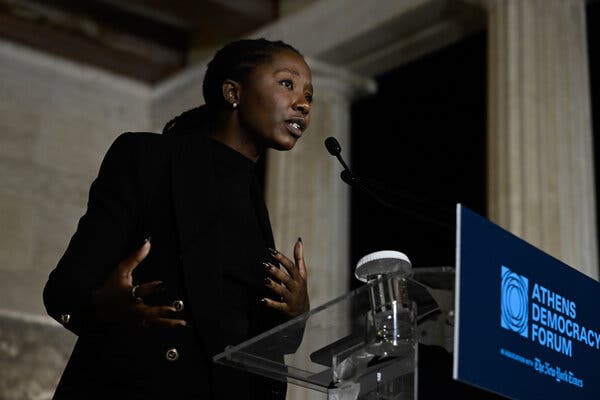 Namatai Kwekweza gestures while speaking into a microphone on a lectern. Gray stone walls and columns stand behind her.