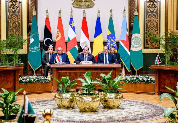 A group of men in suits and ties sitting behind a desk with various flags in the background.