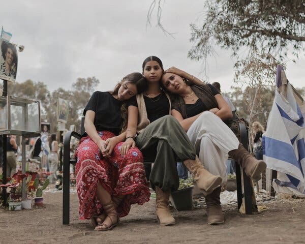 Three women leaning against one another on a bench, with flags and memorials around them and a crowd in the background.