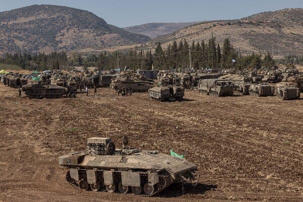 Military vehicles in a dusty landscape with a tree line and mountains in the background.