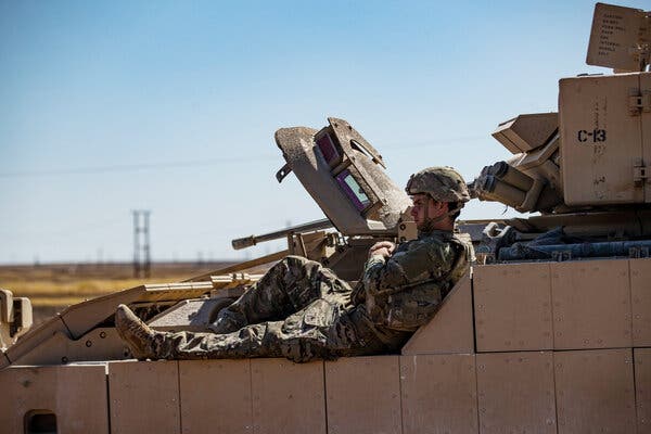 A person in camouflage clothing and helmet sits, leaning back on a brown military vehicle.